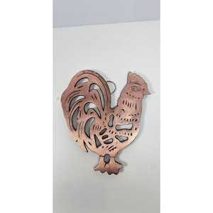 May include: A copper-colored metal rooster-shaped wall hanging. The rooster has a detailed design with cut-out sections in the tail and body. It features a loop at the top for hanging. The metal has a slightly aged appearance.