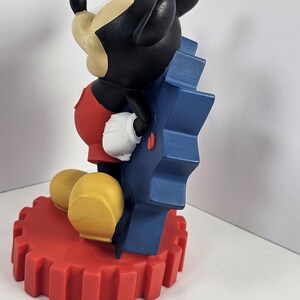 K&ouml;nnte beinhalten: Mickey Mouse Figur, sitzend, mit schwarzem Kopf, wei&szlig;en Handschuhen und gelben Schuhen. Die Figur sitzt auf einem roten Zahnradsockel und lehnt sich an ein blaues Zahnrad.