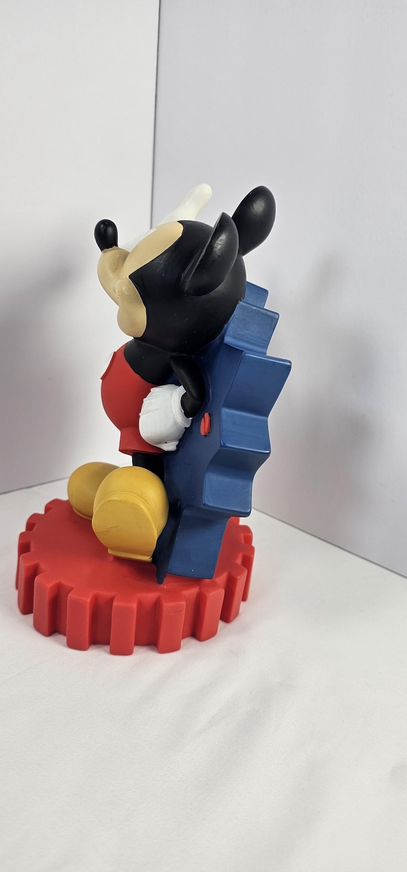 K&ouml;nnte beinhalten: Mickey Mouse Figur, sitzend, mit schwarzem Kopf, wei&szlig;en Handschuhen und gelben Schuhen. Die Figur sitzt auf einem roten Zahnradsockel und lehnt sich an ein blaues Zahnrad.