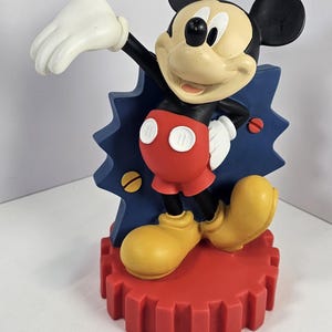 K&ouml;nnte beinhalten: Eine Mickey Mouse-Figur in dynamischer Pose. Die Figur hat einen schwarzen K&ouml;rper, wei&szlig;e Handschuhe, rote Shorts und gelbe Schuhe. Sie steht auf einem roten, zahnradf&ouml;rmigen Sockel mit blauem, blitzf&ouml;rmigem Hintergrund.