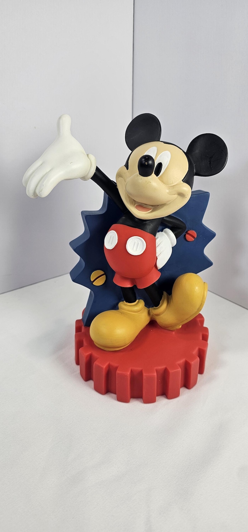 K&ouml;nnte beinhalten: Eine Mickey Mouse-Figur in dynamischer Pose. Die Figur hat einen schwarzen K&ouml;rper, wei&szlig;e Handschuhe, rote Shorts und gelbe Schuhe. Sie steht auf einem roten, zahnradf&ouml;rmigen Sockel mit blauem, blitzf&ouml;rmigem Hintergrund.