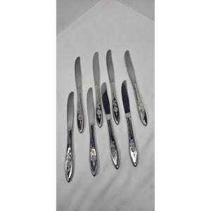 May include: A set of eight silver-colored butter knives with a floral design on the handles. The knives are arranged on a white surface, showcasing their polished finish and decorative details. The blades are curved and the handles are elongated.