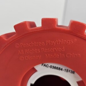 K&ouml;nnte beinhalten: Ein rotes Kunststoff-Spielzeugteil mit wei&szlig;em Innenraum. Die Oberseite hat ein zahnrad&auml;hnliches Design. Text enth&auml;lt "Peachtree Playthings", "All Rights Reserved", "Disney", "Made in China" und einen Produktcode.