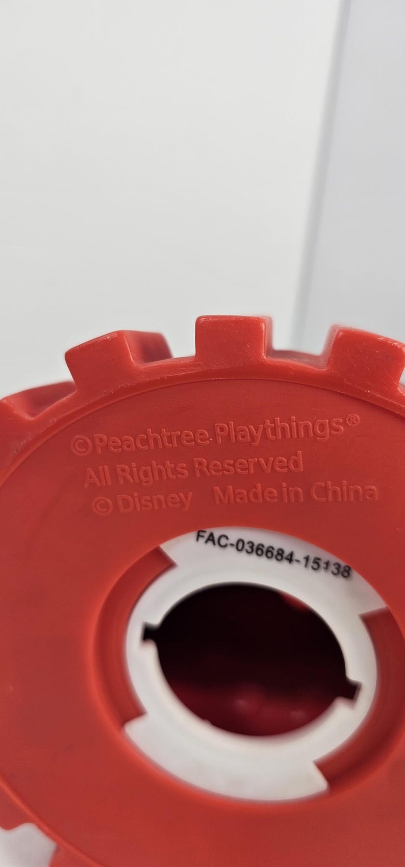 K&ouml;nnte beinhalten: Ein rotes Kunststoff-Spielzeugteil mit wei&szlig;em Innenraum. Die Oberseite hat ein zahnrad&auml;hnliches Design. Text enth&auml;lt "Peachtree Playthings", "All Rights Reserved", "Disney", "Made in China" und einen Produktcode.