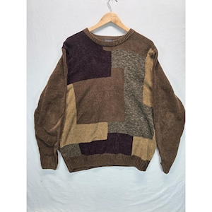 May include: A long-sleeve crew neck sweater featuring a patchwork design of various brown, tan, and dark charcoal gray knit textures. The garment has a ribbed collar, cuffs, and hem, with solid brown sleeves and a multi-paneled front pattern.
