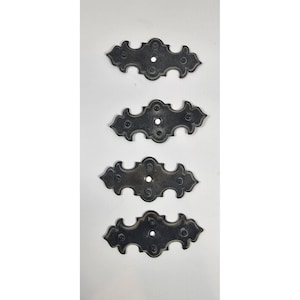 May include: Four antique-style, dark metal decorative plates. Each plate has a symmetrical, ornate design with rounded edges and a central hole, likely for mounting. The plates are arranged vertically against a white background.