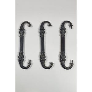 May include: Three antique-style, dark bronze metal hooks. Each hook has a curved top and bottom with a decorative, vertical center featuring heart-shaped and floral designs. The hooks are displayed against a white background.