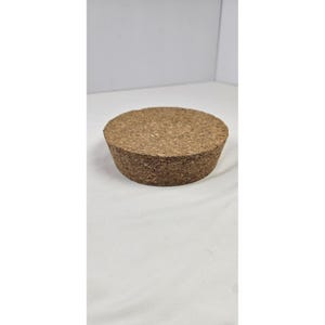 Könnte beinhalten: Ein zylindrischer Korken mit natürlicher brauner Farbe und strukturierter Oberfläche. Der Stopfen hat eine breite, flache Oberseite und einen leicht konischen Körper. Dieser Artikel wird wahrscheinlich zum Verschließen von Flaschen oder Behältern verwendet.