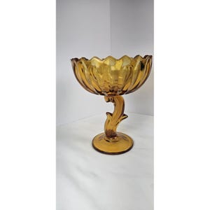 May include: Amber-colored glass pedestal bowl with a scalloped rim. The bowl is supported by a stem with a floral design, and a round base. The glass has a glossy finish and is likely vintage.