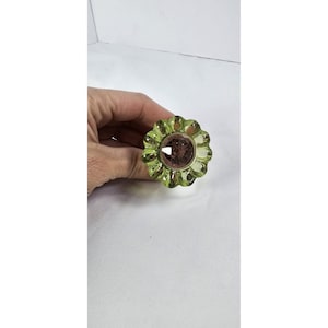 May include: A decorative glass knob with a flower design. The knob features a light green, scalloped edge surrounding a central, faceted, dark pink gem. The knob is held in a hand against a white background.