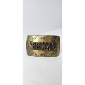 Puede incluir: Hebilla de cinturón rectangular de latón antiguo con la palabra "TEXAS" en letras en relieve. La hebilla tiene un diseño floral ornamentado alrededor de los bordes y las letras.