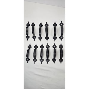 Puede incluir: Un conjunto de doce manijas de puerta de hierro forjado negro. Cada manija tiene un agarre curvo y elementos decorativos en ambos extremos. Las manijas están dispuestas en dos filas de seis, sobre un fondo blanco.