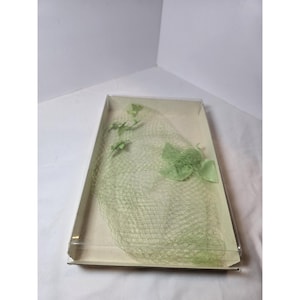 May include: A light green netting veil featuring small fabric floral accents and a larger bow detail, presented inside a rectangular clear plastic box. The delicate mesh fabric is arranged in a soft, gathered shape within the container.