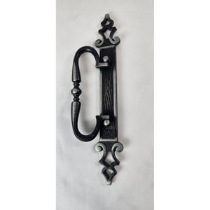 May include: A decorative, antique-style door handle with a curved pull and ornate detailing. The handle is dark gray with silver accents, featuring a vertical plate with a wood grain texture. The design includes decorative flourishes at the top and bottom.