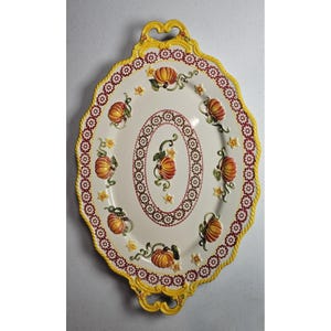 May include: An oval ceramic serving platter with a cream surface, decorated with orange pumpkins, green vines, and yellow flowers. The rim has a yellow rope design and a red floral border. The platter has yellow handles.