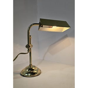 May include: A vintage brass desk lamp with a rectangular shade. The lamp has an adjustable arm and a round base. The shade is a muted green color, and the lamp is lit, casting a warm glow. The lamp is a classic design.