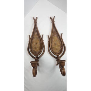 May include: Pair of vintage wall sconces. Each sconce features a brown, tulip-shaped frame with a woven cane center. A small cup-shaped holder is positioned below the cane, and a curved, decorative element extends downward. These sconces are ideal for adding a touch of vintage charm to any space.
