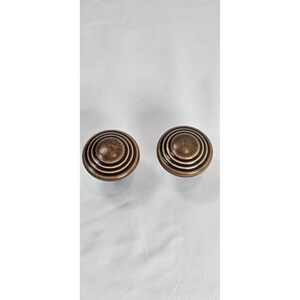 May include: Two bronze cabinet knobs with a concentric circle design. The knobs have a rounded shape and a slightly aged appearance, suggesting a vintage or antique style. They are likely used for furniture or cabinetry.