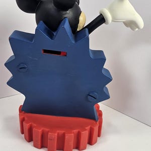 K&ouml;nnte beinhalten: Eine Mickey Mouse-Sparb&uuml;chse. Die Bank hat einen schwarzen Mickey Mouse-Kopf, einen blauen sternf&ouml;rmigen K&ouml;rper und eine rote zahnradf&ouml;rmige Basis. Mickeys wei&szlig;e behandschuhte Hand ist zum Gru&szlig; erhoben.