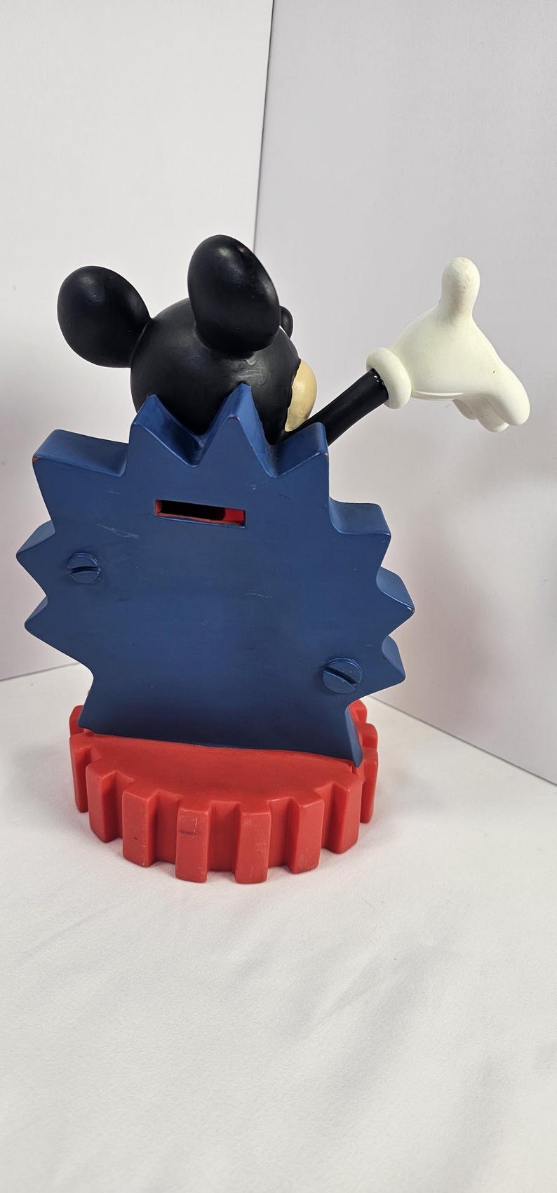 K&ouml;nnte beinhalten: Eine Mickey Mouse-Sparb&uuml;chse. Die Bank hat einen schwarzen Mickey Mouse-Kopf, einen blauen sternf&ouml;rmigen K&ouml;rper und eine rote zahnradf&ouml;rmige Basis. Mickeys wei&szlig;e behandschuhte Hand ist zum Gru&szlig; erhoben.