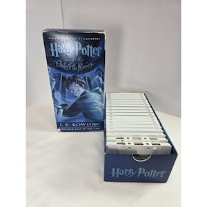 May include: A Harry Potter and the Order of the Phoenix audiobook set featuring a blue box with an illustration of a character with glasses and the text J.K. Rowling and Jim Dale. The set includes seventeen white cassette tapes in a blue holder.