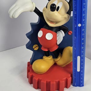 K&ouml;nnte beinhalten: Mickey Mouse Figur, mit schwarzem Kopf und Ohren, beigem Gesicht und roter Hose. Die Figur steht auf einem roten, zahnradf&ouml;rmigen Sockel und ist etwa 25 cm hoch. Der Hintergrund enth&auml;lt ein blaues Dekorationselement.