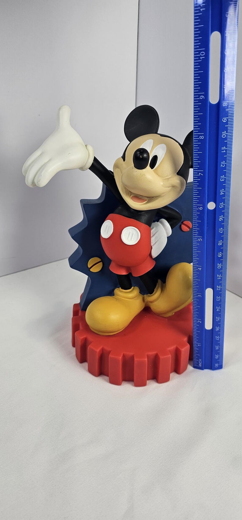 K&ouml;nnte beinhalten: Mickey Mouse Figur, mit schwarzem Kopf und Ohren, beigem Gesicht und roter Hose. Die Figur steht auf einem roten, zahnradf&ouml;rmigen Sockel und ist etwa 25 cm hoch. Der Hintergrund enth&auml;lt ein blaues Dekorationselement.