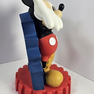 K&ouml;nnte beinhalten: Eine Mickey Mouse-Figur in klassischer Pose. Die Figur ist haupts&auml;chlich schwarz, mit roten Shorts, gelben Schuhen und wei&szlig;en Handschuhen. Sie steht auf einem roten, zahnradf&ouml;rmigen Sockel mit blauem Akzent.