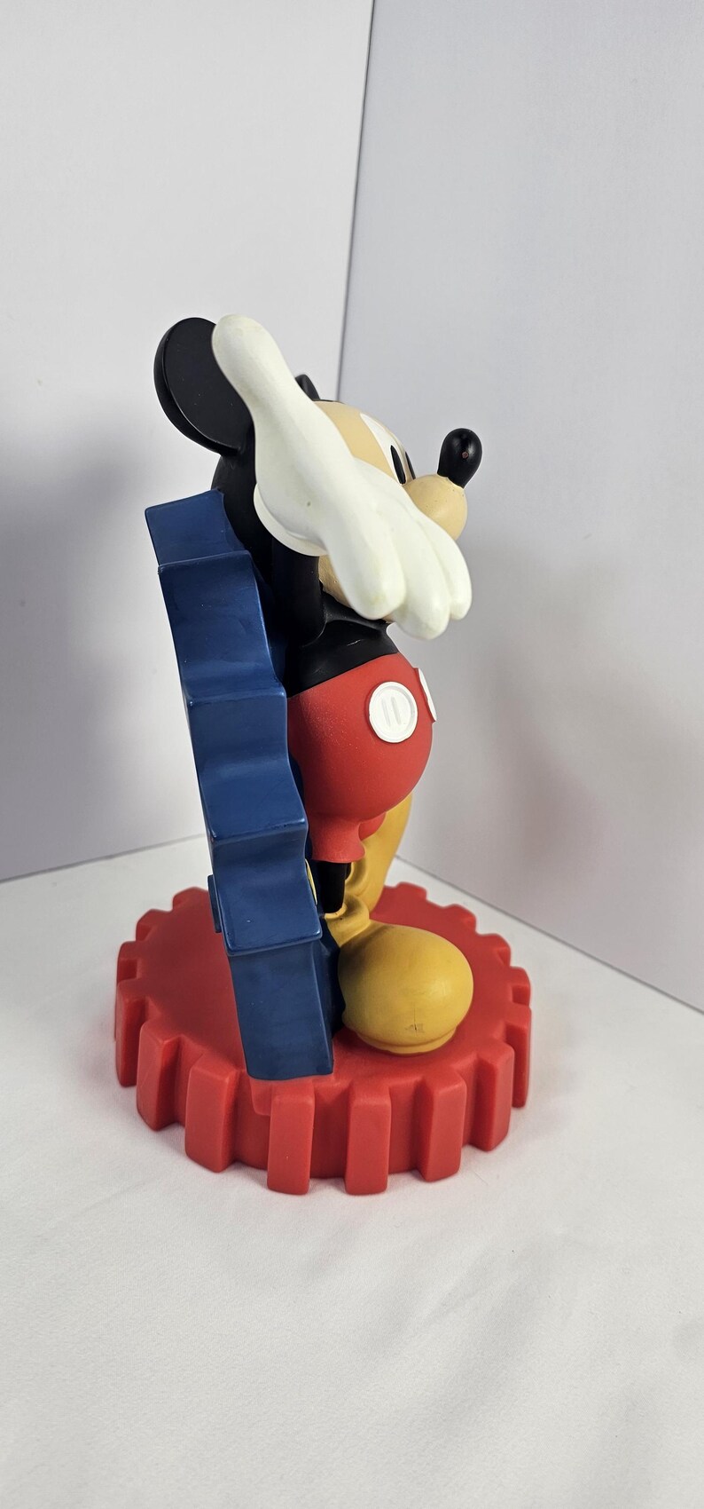 K&ouml;nnte beinhalten: Eine Mickey Mouse-Figur in klassischer Pose. Die Figur ist haupts&auml;chlich schwarz, mit roten Shorts, gelben Schuhen und wei&szlig;en Handschuhen. Sie steht auf einem roten, zahnradf&ouml;rmigen Sockel mit blauem Akzent.