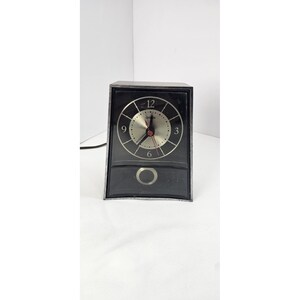May include: Black vintage clock with a silver face and hands. The clock has a rectangular shape with a silver trim and a small drawer at the bottom. The clock face features the numbers 12, 3, 6, and 9.
