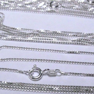 May include: A close-up of multiple silver box chains of varying lengths. The chains are arranged in a layered pattern, with the shortest chain at the top and the longest chain at the bottom. The chains are all made of a fine, delicate metal.