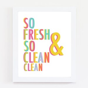May include: A framed print featuring the words "SO FRESH SO CLEAN & CLEAN" in vibrant, layered block letters. The text uses pink, green, yellow, and blue against a white backdrop. The print is encased in a white frame.