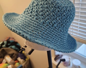 May include: A light blue crochet hat with a wide brim and a slightly conical crown. The hat is made of a textured, woven material. The hat is displayed on a white mannequin head, mounted on a black tripod.