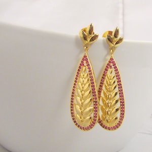 May include: A pair of gold dangle earrings with a teardrop shape. The earrings are decorated with a leaf design and set with small pink gemstones.