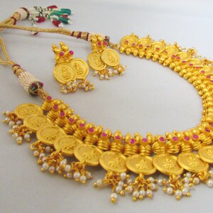 May include: A gold necklace with a coin design and pink accents. The necklace has a matching pair of earrings with a similar design. The necklace is made of gold-plated metal and features a series of round coins with a raised design. The coins are connected by a chain and have small pink stones set in the center of each coin. The earrings are also made of gold-plated metal and feature a single coin with a raised design and a pink stone in the center. The earrings are attached to a small hook.