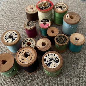 May include: A collection of vintage wooden spools of thread in various colors, including green, blue, and red. Some spools have labels with text. The spools are arranged on a neutral-colored surface, showcasing a variety of thread types.