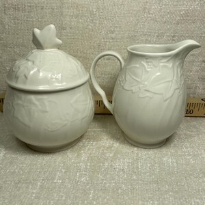 May include: A white ceramic sugar bowl with a lid and a matching creamer. Both pieces feature a raised leaf design. The sugar bowl has a leaf-shaped handle on the lid. The creamer has a curved handle and a spout.