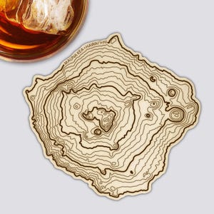 Könnte beinhalten: Ein Untersetzer aus Holz mit einem topografischen Kartendesign. Der Untersetzer zeigt Konturlinien und den Text "MAUNA KEA HAWAII HI" in einem kreisförmigen Muster. Der Untersetzer ist hellbraun mit dunkelbraunen Linien und Text.
