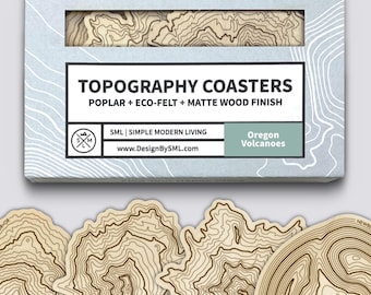 May include: A set of topography coasters made of poplar, eco-felt, and matte wood finish. The coasters feature a detailed contour map design in shades of brown and beige. The packaging includes the text "TOPOGRAPHY COASTERS" and "Oregon Volcanoes".