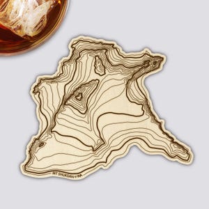 Könnte beinhalten: Ein hellfarbiger Untersetzer mit einem topografischen Kartendesign des Mt. Shuksan, WA, mit braunen Konturlinien. Der Untersetzer hat eine einzigartige Form und der Text "MT SHUKSAN WA" ist unten aufgedruckt. Ein Glas bernsteinfarbener Flüssigkeit mit Eis befindet sich in der oberen linken Ecke.