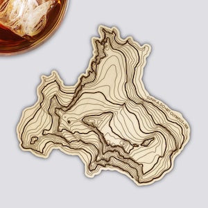 Könnte beinhalten: Ein Untersetzer aus Holz mit einem topografischen Kartendesign. Der Untersetzer hat Konturlinien in Dunkelbraun auf hellbraunem Holzmaserung. Der Text "SUNH-A-DO MT OLYMPUS WA" ist aufgedruckt.