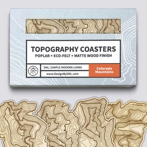 Könnte beinhalten: Ein Set aus Holz-Topographie-Untersetzern mit mattem Holzfinish. Die Untersetzer bestehen aus Pappel und Öko-Filz. Auf der Box steht "TOPOGRAPHY COASTERS" und "Colorado Mountains". Die Untersetzer haben ein detailliertes Kartendesign.