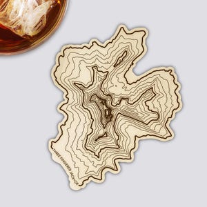 Könnte beinhalten: Ein Untersetzer aus Holz mit einem topografischen Kartendesign mit braunen Konturlinien. Der Untersetzer hat die Form einer Landmasse und trägt den Text "THREE FINGERED JACK OR". Ein Glas bernsteinfarbener Flüssigkeit mit Eis befindet sich oben links.