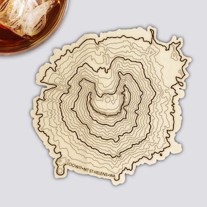 Könnte beinhalten: Ein hellfarbiger Holzuntersetzer mit einem topografischen Kartendesign des Mount St. Helens, Washington. Der Untersetzer zeigt Konturlinien in Dunkelbraun und den Text "LOOWIT MT ST HELENS WA". Ein Glas bernsteinfarbener Flüssigkeit mit Eis befindet sich oben links.