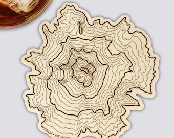 May include: A light-colored coaster with a topographic map design of Mt. Hood, Oregon. The coaster has dark brown contour lines and the text "WY'EAST • MT HOOD • OR" at the bottom. A glass of amber liquid with ice cubes is in the upper left corner.