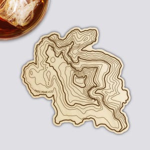 Könnte beinhalten: Ein Holzuntersetzer mit einem topografischen Kartendesign des Pikes Peak, mit Konturlinien in Dunkelbraun auf hellem Holzgrund. Der Untersetzer hat eine unregelmäßige Form und trägt den Text "PIKES PEAK + CO". Ein Glas bernsteinfarbener Flüssigkeit mit Eis ist teilweise sichtbar.