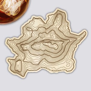 Könnte beinhalten: Ein hellfarbiger Untersetzer mit einem topografischen Kartendesign von Mammoth Mountain, CA. Der Untersetzer hat braune Konturlinien und den Text "Mammoth Mountain CA". Ein Glas bernsteinfarbener Flüssigkeit mit Eis ist oben links zu sehen.