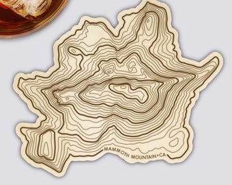 May include: A light-colored coaster with a topographic map design of Mammoth Mountain, CA. The coaster features brown contour lines and the text "Mammoth Mountain CA" at the bottom. A glass of amber liquid with ice is visible in the upper left corner.