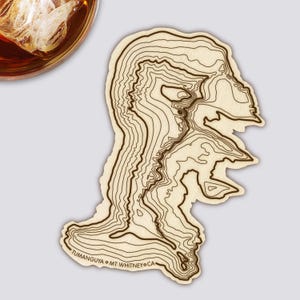 Könnte beinhalten: Ein Untersetzer aus Holz mit einem topografischen Kartendesign. Der Untersetzer hat braune Konturlinien auf hellem Holzgrund. Der Text "TUMANGUYA • MT WHITNEY CA" ist am unteren Rand aufgedruckt. Ein Glas bernsteinfarbener Flüssigkeit mit Eis befindet sich oben links.