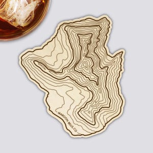 Könnte beinhalten: Holzuntersetzer mit einem topografischen Kartendesign von Maroon Bells, CO. Der Untersetzer ist hellbraun mit dunkelbraunen Konturlinien. Ein Glas bernsteinfarbener Flüssigkeit mit Eis befindet sich in der oberen linken Ecke.
